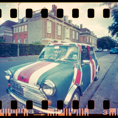 The image is a black and white photo of an old-fashioned car parked on the side of a street. The car has a red, white, and blue color scheme with a Union Jack design. It appears to be a vintage model, possibly a Volkswagen Beetle.