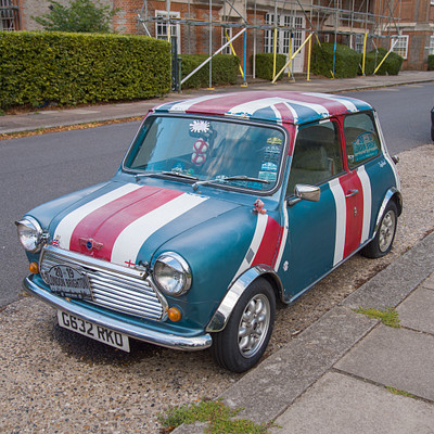 The image features a blue and red striped car parked on the side of a street. It is an old-fashioned model, possibly a Volkswagen Beetle, with a distinctive British flag design painted on its hood. The car is parked next to a curb, and there are several other cars in the vicinity, including one behind it and another further down the road.