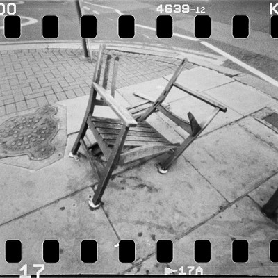 The image is a black and white photo of an outdoor scene featuring a wooden chair that has been turned upside down. The chair is placed on the sidewalk, possibly as part of an art installation or simply discarded. There are several benches in the background, with one located near the center of the scene and another towards the right side.