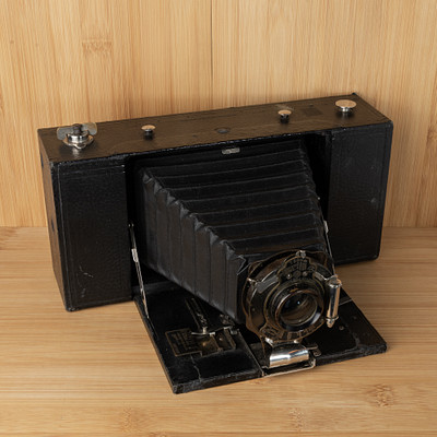 The image features an old-fashioned camera sitting on a wooden table. It is placed in the center of the scene, drawing attention to its vintage design. The camera appears to be black and has a leather texture, giving it a classic appearance.