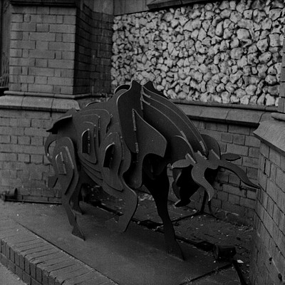 The image is a black and white photo of an animal statue, possibly a bull or a horse. It appears to be made from metal and has been placed on a brick walkway. The statue is positioned in front of a wall with a pile of rocks behind it.