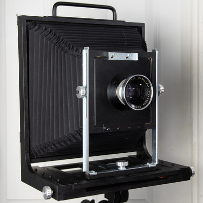 The image features an old-fashioned camera with a black case, sitting on top of a stand. It appears to be an antique or vintage model, possibly a Polaroid camera. The camera is placed against a wall, and the stand it's resting on has a metal base.