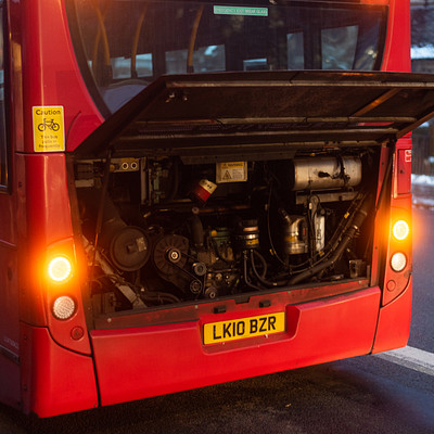 The image features a red bus with its hood open, revealing the engine and various mechanical parts. The bus is parked on a street, possibly in a parking lot or near a building. There are two people visible in the scene, one standing closer to the left side of the bus and another person further back on the right side.