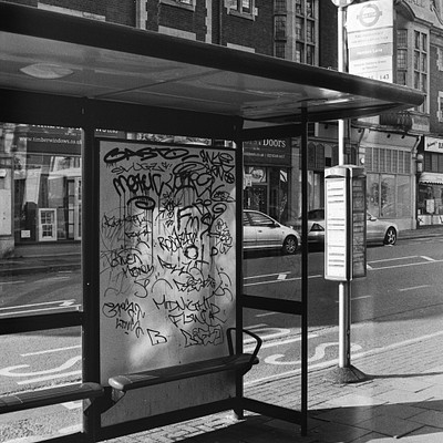 The image is a black and white photo of a bus stop with a bench in front of it. There are several cars parked nearby, including one on the left side of the scene, two behind the bench, and another further back to the right. A traffic light can be seen above the street, indicating that this is an urban setting.