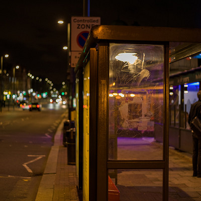 The image is a black and white photo of a man standing outside of a telephone booth at night. He appears to be waiting for someone or something, possibly a taxi or another person. There are several cars parked in the background, with one car on the left side of the scene and two others further back on the right side.