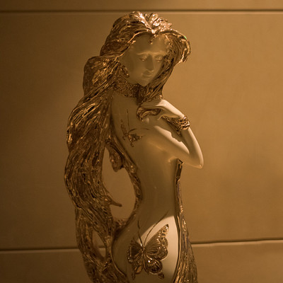 The image features a statue of a woman with long hair, wearing a gold dress. She appears to be naked and has an intricate design on her body. The statue is positioned in front of a wall, which adds depth to the scene.