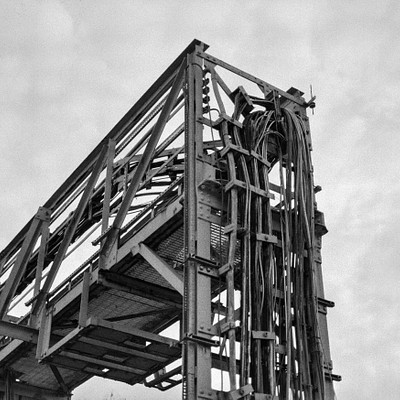 The image is a black and white photo of an old, rusted metal structure. It appears to be a large piece of machinery or a bridge with a staircase leading upwards. There are several people in the scene, some standing on the stairs while others are scattered around the area.