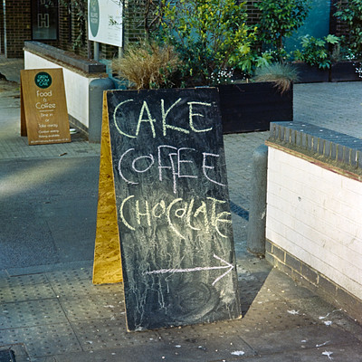 The image features a black chalkboard sign on the sidewalk, advertising cake and coffee. The sign is placed in front of a building with a brick facade. There are several potted plants surrounding the area, adding to the ambiance.