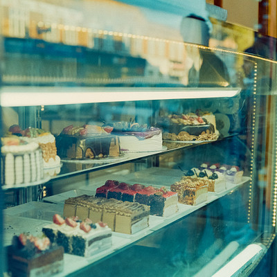 The image features a display case filled with an assortment of cakes and pastries. There are at least 13 different types of cakes visible in the case, each with unique designs and flavors. Some of these cakes are placed on trays or plates, while others are arranged directly within the glass case.