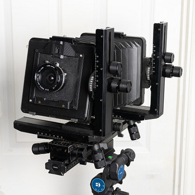 The image features a black camera mounted on a tripod, which is placed against a white wall. The camera appears to be an old-fashioned model with a vintage look. There are several buttons and knobs visible on the camera, indicating its functionality and adjustability.