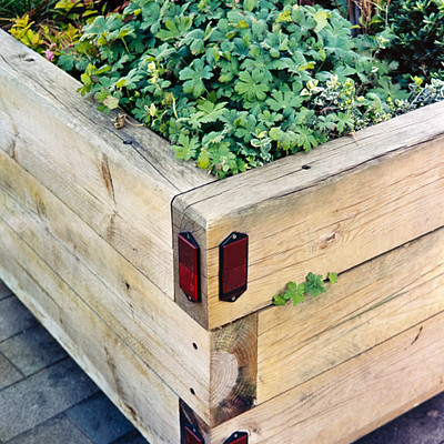 The image features a wooden planter box filled with various plants and flowers. There are several potted plants placed inside the box, creating an attractive garden display. Some of these plants have red stakes in them, adding a pop of color to the scene.