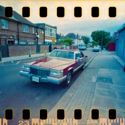 The image is a vintage photo of an old car parked on the side of a street. It appears to be a red or maroon color, and it has a chrome grill. There are several other cars in the scene, including one behind the main car and two others further down the road.