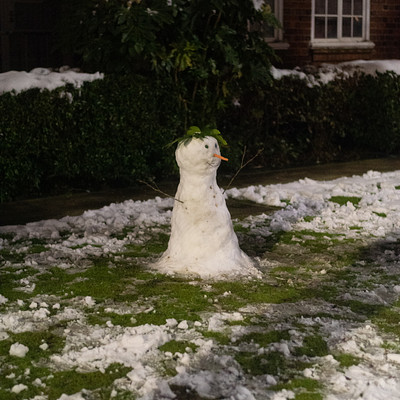 The image features a snowman sitting in the middle of a yard. It is surrounded by a green lawn and has a brick house nearby. There are several windows on the house, with some located near the top left corner and others towards the right side.
