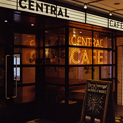 The image is a black and white photo of the exterior of a restaurant called Central Cafe. The cafe has a neon sign on its door, making it stand out in the nighttime setting. There are several chairs placed outside the entrance, likely for customers to sit while waiting or enjoying their meals.