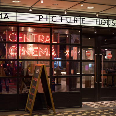 The image features a movie theater with its entrance door open, inviting people to enter. A large neon sign is prominently displayed above the entrance, advertising the Central Cinema Picture House. Inside the theater, there are several chairs arranged in rows for patrons to sit and enjoy their movies.