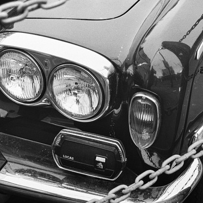 The image is a black and white photo of an old car with its hood open. The car appears to be parked on the street, possibly in front of a building or near some chains. The car's headlights are visible, adding to the vintage feel of the scene.