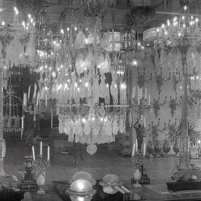 The image is a black and white photograph of an ornate chandelier hanging from the ceiling. The chandelier features numerous lights, creating a dramatic effect in the room. There are also several other chandeliers visible throughout the space, adding to the overall grandeur of the scene.