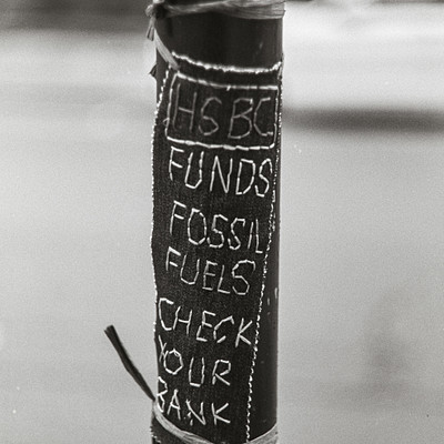 The image is a black and white photo of a street pole with a sign attached to it. The sign reads "Fossil Fuels Check Your Bank." Above the sign, there are two cars parked on the side of the road. One car is positioned further back in the scene, while the other car is closer to the foreground.