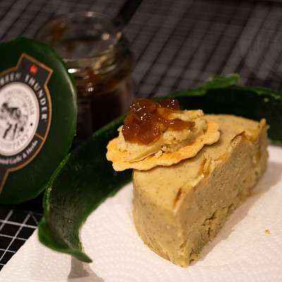 The image features a slice of cake sitting on top of a white plate. The cake is placed in the center of the plate, and it appears to be a dessert or snack item. Next to the cake, there are two jars with lids, one containing jelly and the other possibly holding some sauce.