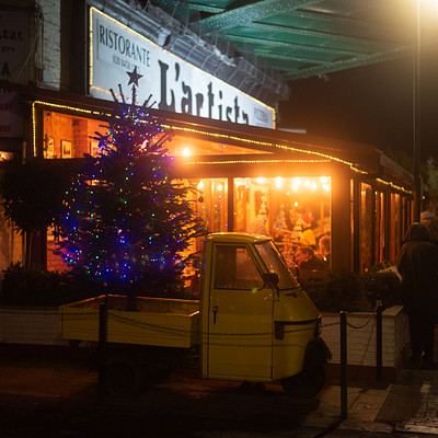 The image features a small yellow truck parked in front of a restaurant. The truck is positioned under an illuminated Christmas tree, creating a festive atmosphere. There are several people around the area, some standing near the truck and others walking by.
