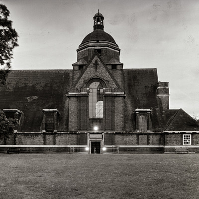 The image is a black and white photo of an old church with a large steeple. The building has a brick exterior, giving it a classic appearance. There are two windows on the front of the church, one larger than the other.