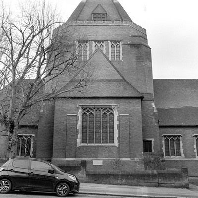 The image is a black and white photo of an old church with a brick exterior. A small car is parked in front of the church, near the left side of the building. There are several windows on the church, some of which have arches above them.