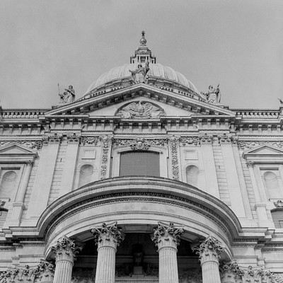 The image is a black and white photo of an old building with a large dome on top. The building has many statues, including one in the center of the front facade. There are also two smaller statues located near the bottom left corner of the building.