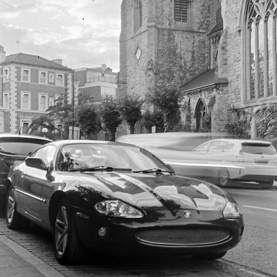The image is a black and white photo of a city street with several cars parked on the side. A sports car, possibly a Corvette, is prominently featured in the foreground, occupying most of the frame. Other cars are visible in various positions along the street, including one behind the sports car and others further down the road.