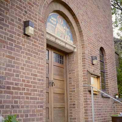 The image features a large brick building with an arched doorway. Above the door, there is a stained glass window depicting Jesus and Saint Philip. The doorway has a wooden frame and appears to be made of wood as well.