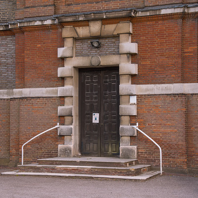 The image features a large brick building with an arched doorway. Above the door, there is a clock mounted on the wall. The doorway has a metal railing in front of it, providing safety and accessibility for those entering or exiting the building.