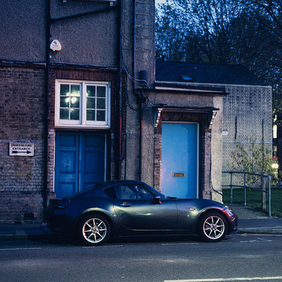 The image features a black sports car parked on the side of a street, in front of a blue door. The car is positioned close to a building and appears to be parked next to a house. There are several other cars visible in the scene, with one located further down the street and two others parked closer together.
