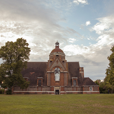 The image features a large brick building with a steeple, which appears to be an old church. The building is surrounded by trees and has a grassy area in front of it. There are two people standing near the building, possibly admiring its architecture or attending an event.