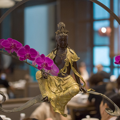 The image features a statue of Buddha sitting on a chair, surrounded by purple flowers. The statue is positioned in the center of the scene and appears to be made of gold or bronze. There are several people in the background, some seated at dining tables with cups and bowls placed on them.