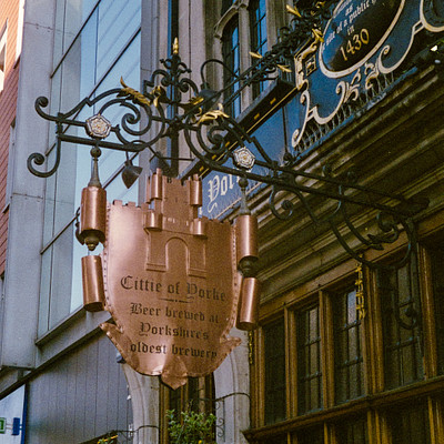 The image features a large, ornate sign hanging from the side of an old building. The sign is made up of various pieces and has a metal design. It appears to be a street sign or a decorative piece attached to the building.