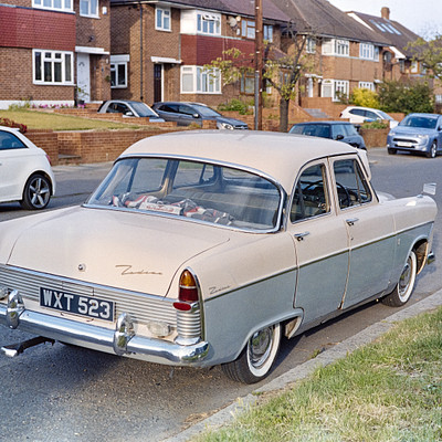 The image features a vintage car parked on the side of a street. It is an old-fashioned vehicle with a tan color, and it appears to be a classic model. There are several other cars in the scene, including one behind the vintage car and others parked further down the road.