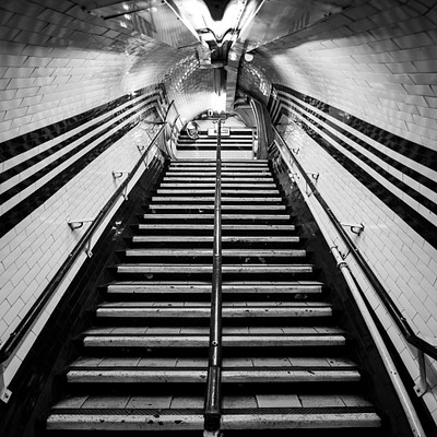 The image is a black and white photo of an old staircase, likely in a subway or train station. There are several steps leading upwards, with the first step being closer to the bottom left corner of the frame. A person can be seen standing at the top of the stairs, possibly waiting for a train or preparing to descend.