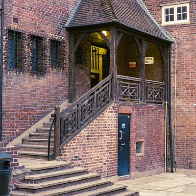 The image features a brick building with an old-fashioned design, including a steep staircase leading to the entrance. Above the doorway, there is a small balcony or porch area that adds charm and character to the structure. Inside the building, there are several people visible, likely going about their daily activities.