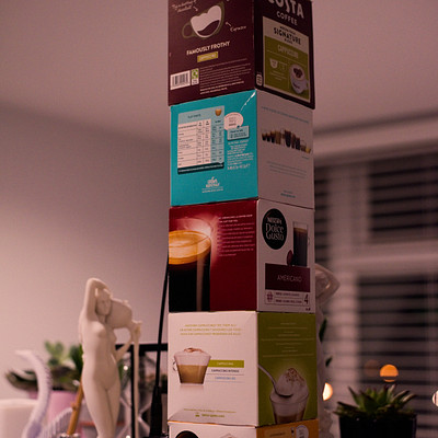 The image features a stack of coffee boxes on top of each other, with the tallest box at the bottom and the smallest one at the top. There are several cups placed around the boxes, some near the top and others closer to the bottom. A potted plant is situated in the background, adding a touch of greenery to the scene.