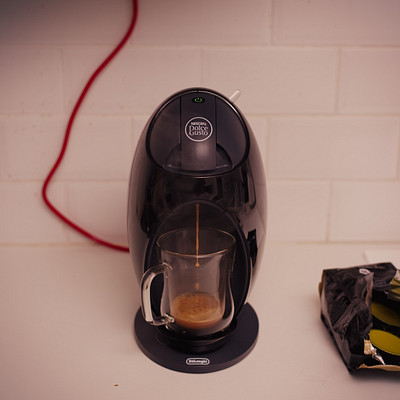 The image features a black coffee maker sitting on top of a white counter. A cup is placed next to the coffee maker, ready for use. There are also two bags of coffee beans nearby, one located closer to the left side and another towards the right side of the scene.