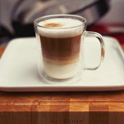 A white plate with a cup of coffee on it. The cup is filled with a mixture of milk and espresso, creating a layered appearance. The cup is placed on top of the plate, which sits on a wooden table.