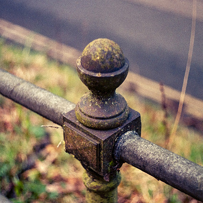 The image features a metal fence with a stone post and a metal ball on top of it. The fence is located near the side of a road, possibly in a park or garden setting. There are also some grassy areas visible around the fence, adding to the natural atmosphere of the scene.