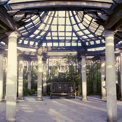 The image features a large, open area with several columns and arches. There is an outdoor seating arrangement in the middle of the space, surrounded by pillars. A bench can be seen near the center of the scene, while another one is located further back on the right side.