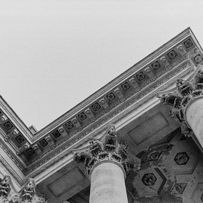 The image is a black and white photo of an old building with columns. There are several statues on the building, including one near the top left corner, another in the middle right area, and two more towards the bottom right side. The architecture appears to be ornate and intricate, showcasing the historical significance of the structure.