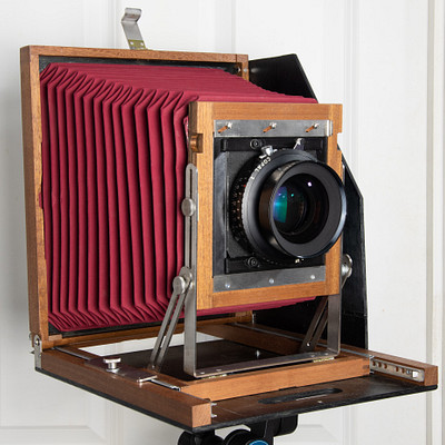 The image features an old-fashioned camera with a red and brown color scheme. It is placed on a wooden stand, which appears to be a tripod. The camera has a large lens in the front, giving it a vintage appearance.