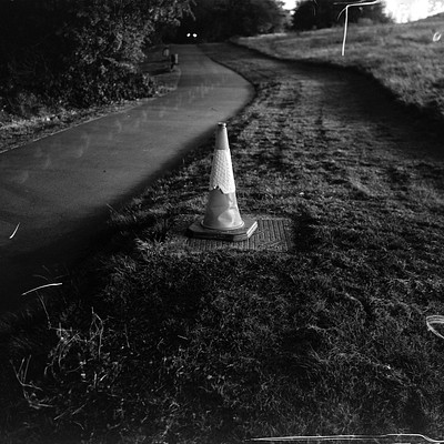 The image is a black and white photo of an orange traffic cone sitting on the side of a road. The cone appears to be placed in the grass, possibly near a street or pathway. There are no other objects or people visible in the scene.