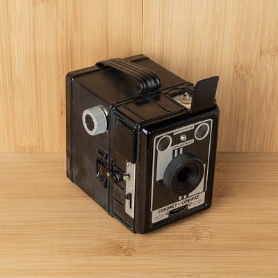 The image features a black and silver camera sitting on top of a wooden table. The camera is an old-fashioned model, possibly a vintage Polaroid or Kodak instant camera. It has a strap attached to it, which could be used for carrying the camera around.