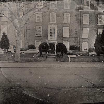 The image is a black and white photo of a street scene with several cars parked along the side. There are two trucks, one on the left side and another further down the road. A few cars can be seen in various positions throughout the scene.