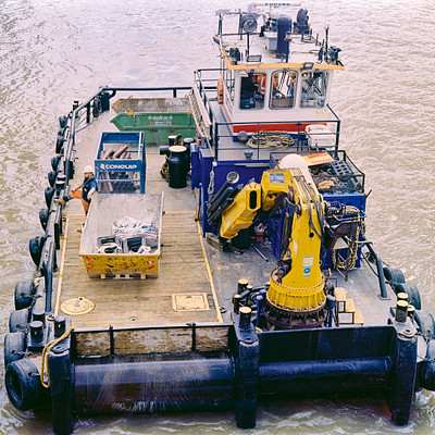 The image features a large boat floating on the water, with a crane and a yellow machine on top of it. There are several people visible around the boat, some standing closer to the edge while others are further away. A person is also wearing an orange vest, which suggests they might be working in a potentially hazardous environment.