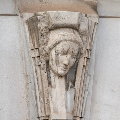The image features a stone carving of a woman's face, which appears to be an artistic decoration. The carved figure is situated on the side of a building and has a unique design that adds character to the structure.