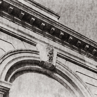 The image is a black and white photo of an old building with a stone archway. Above the arch, there is a carved stone figure that appears to be a lion or a similar animal. The building has many columns and features a large arched doorway.